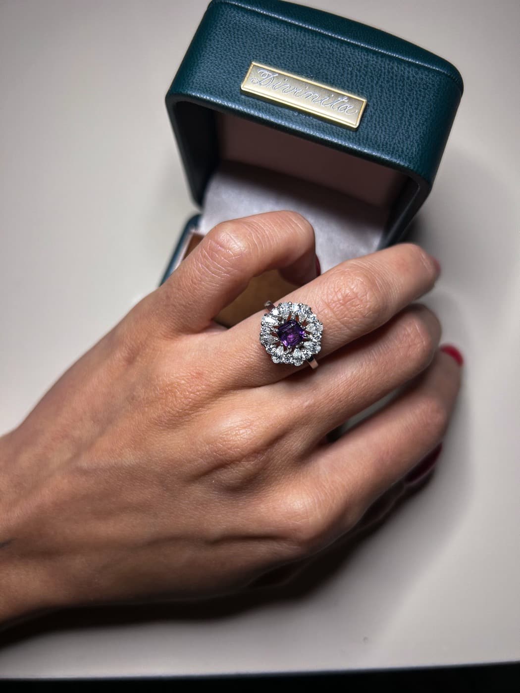 Anel Divinita finalizado com ametista e halo de diamantes, na mão, ao lado do estojo